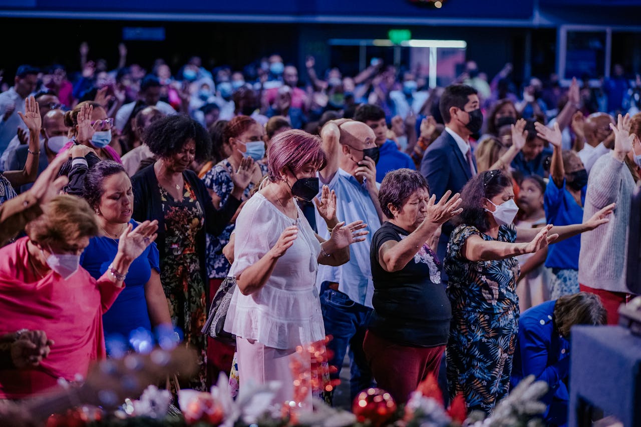 A large, diverse group of people praying and worshipping together indoors, wearing face masks.